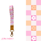 Three colourful fabric wristlet keychains with gold hardware and tan leather ends, featuring pink gingham daisy, blush polka dot stripe, and retro orange checker floral patterns, laid on pastel notebooks with floral accents – Lilly Jayne Collective.