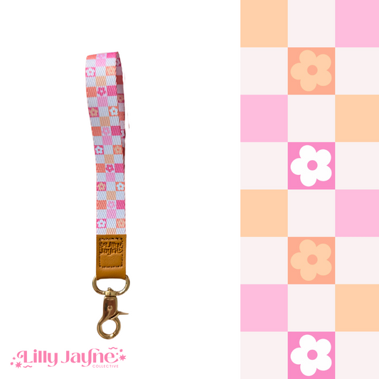 Three colourful fabric wristlet keychains with gold hardware and tan leather ends, featuring pink gingham daisy, blush polka dot stripe, and retro orange checker floral patterns, laid on pastel notebooks with floral accents – Lilly Jayne Collective.