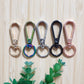 The Coffee Teach Repeat Teacher Lanyard by Lilly Jayne Collective features five metal keychain clasps—gold, rainbow, silver, black, and rose gold—arranged on a wood background with green leaves for a stylish DIY accessory.