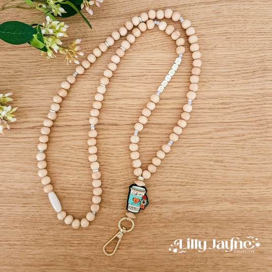 A wooden bead personalized lanyard with a metal clip and a key ring, featuring a breakaway safety clasp, laid out on a wooden surface with decorative flowers in the background.