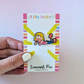 Enamel pin on a card held by a hand with a light purple background