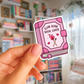 Pink book-shaped sticker with 'Slow Burn Book Lover' text held in front of a bookshelf.