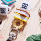The Lilly Jayne Collective Coffee Teach Repeat Teacher Lanyard features charms of stacked books, a sunflower, and a coffee teach repeat mug, displayed on wood with coffee beans, books, and a plant leaf.