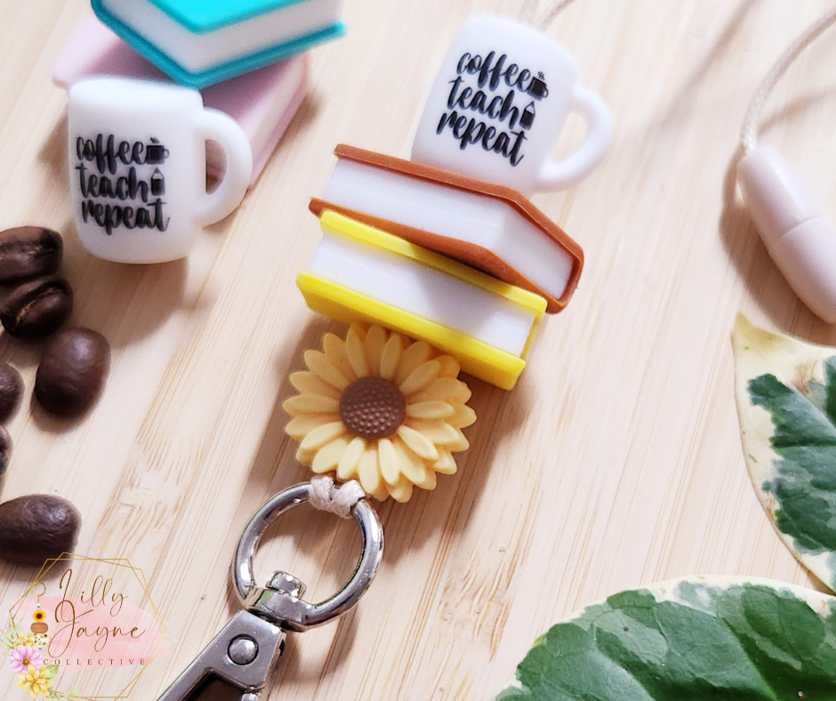 The Lilly Jayne Collective Coffee Teach Repeat Teacher Lanyard features charms of stacked books, a sunflower, and a coffee teach repeat mug, displayed on wood with coffee beans, books, and a plant leaf.