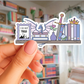 Sticker with dragon, books, and sword held by a hand in front of a bookshelf.