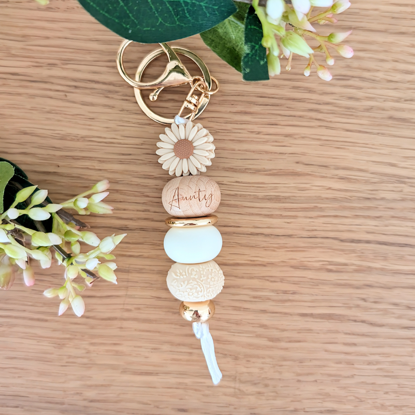 Handmade keyring with beige daisy flower charm, engraved 'Aunty' wooden bead, floral patterned bead, and pastel pink accents on a wooden surface with greenery