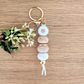 Handmade keyring with white daisy flower charm, engraved 'Aunty' wooden bead, floral patterned bead, and pastel pink accents on a wooden surface with greenery