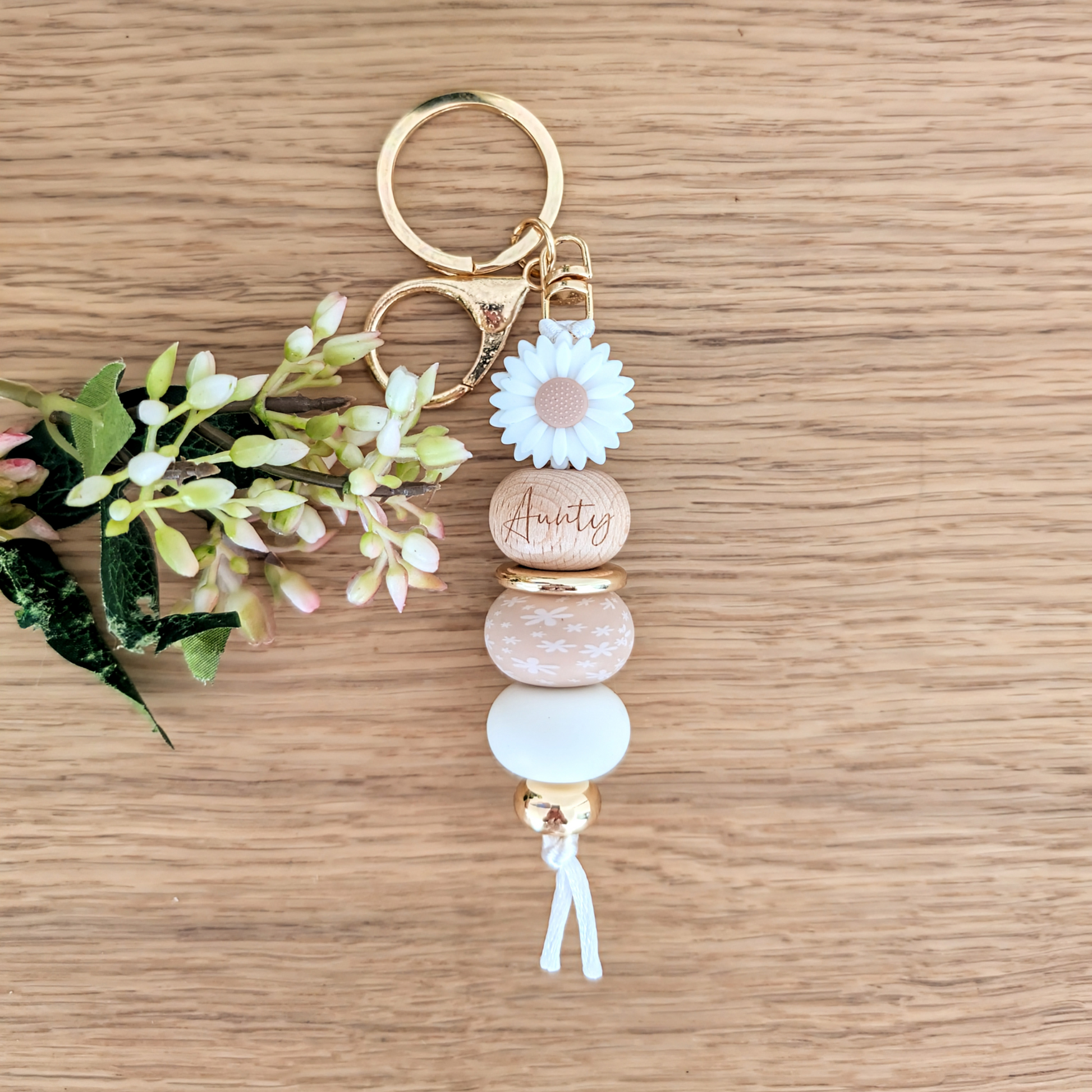 Handmade keyring with white daisy flower charm, engraved 'Aunty' wooden bead, floral patterned bead, and pastel pink accents on a wooden surface with greenery