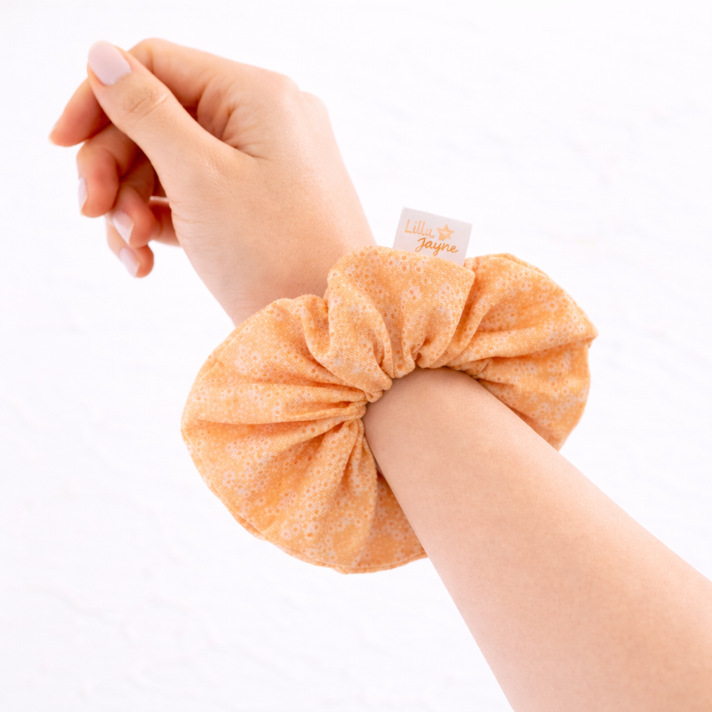 Hand wearing an orange scrunchie with a visible brand label on a white background