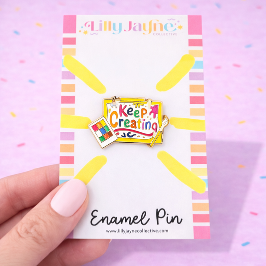 Colorful enamel pin on a card with 'Lilly Jayne Collective' branding, held by a hand against a pink background.