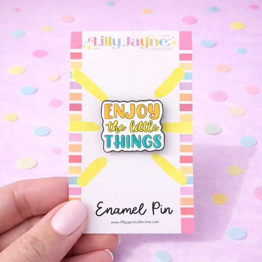 Enamel pin with 'Enjoy the little things' text on a colorful card, held against a pink background with polka dots.