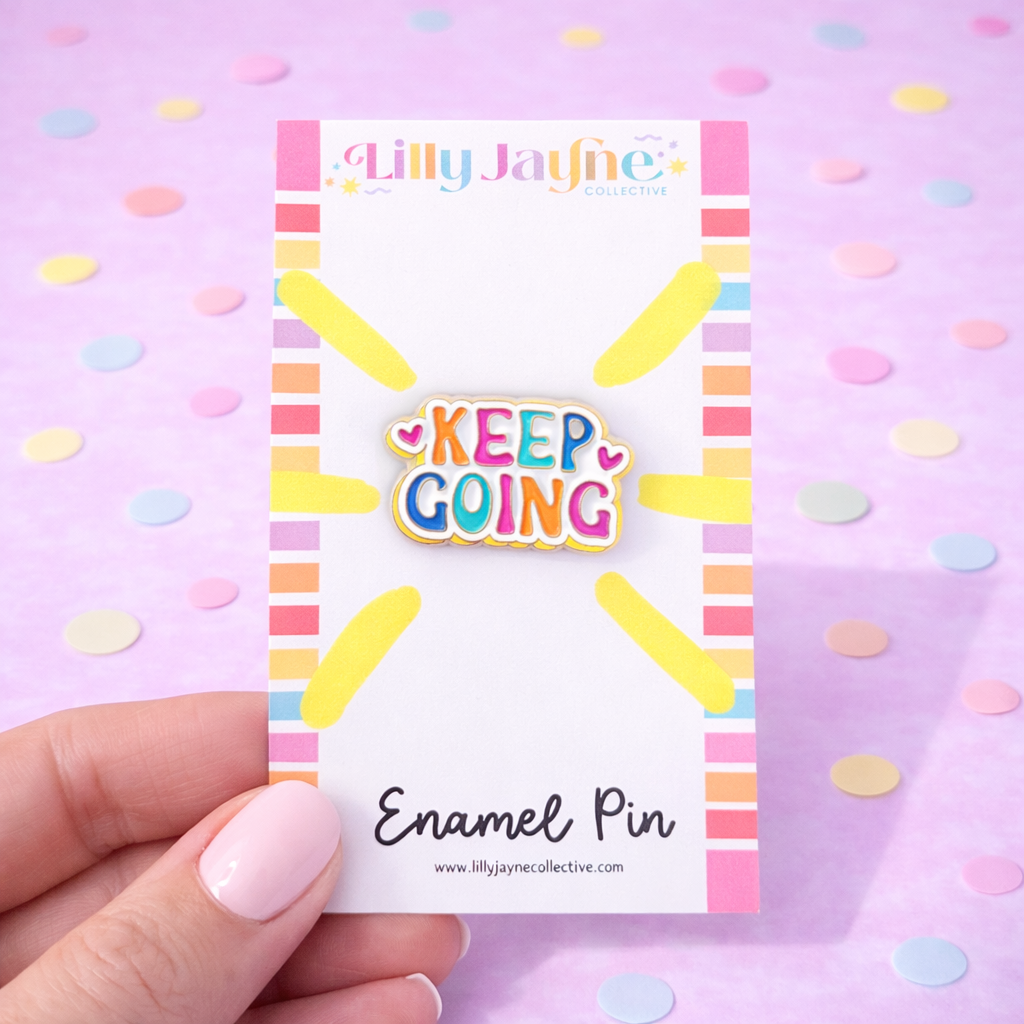 Enamel pin with 'Keep Going' text on a colorful card against a pink background
