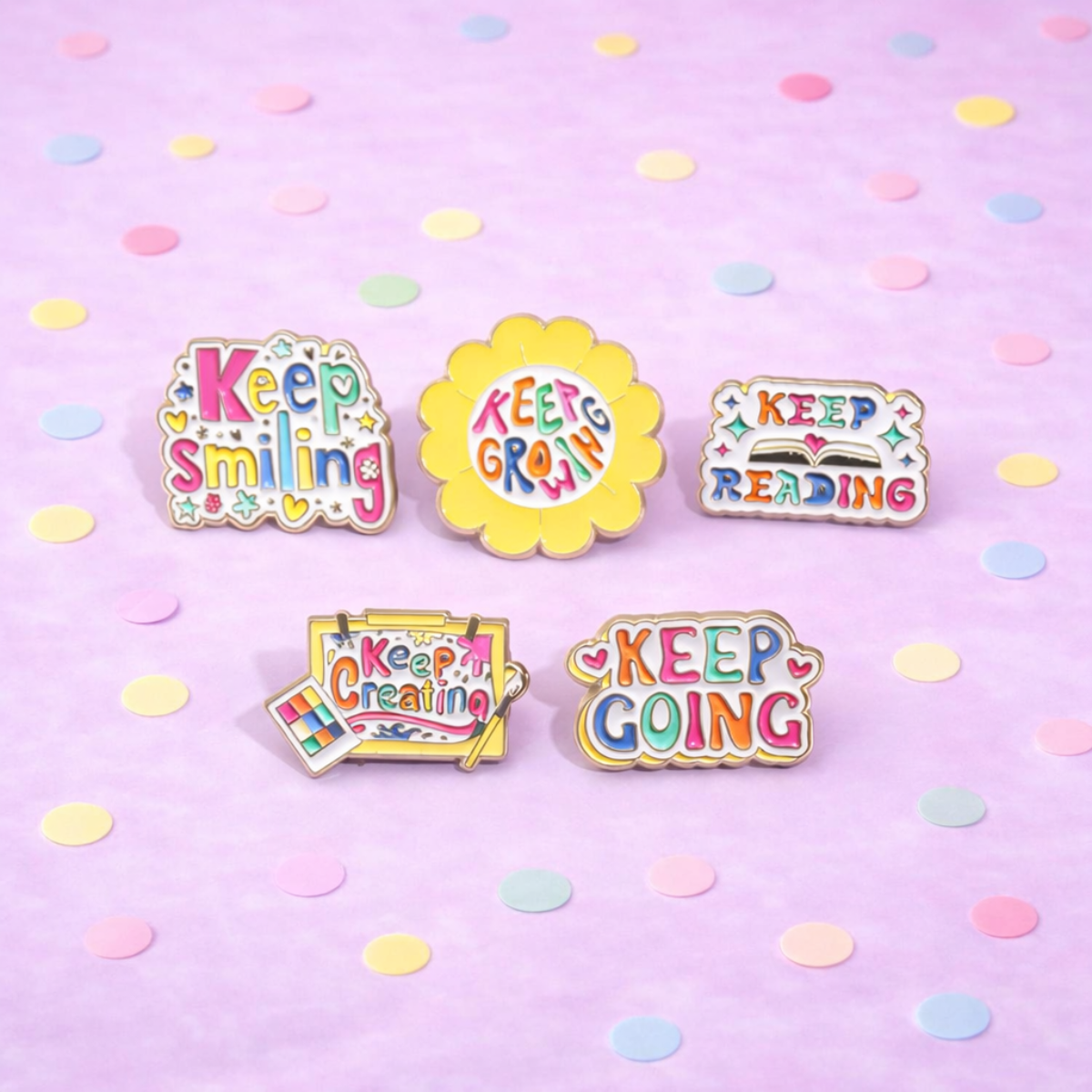 Five colorful enamel pins with motivational phrases on a pink background with confetti.