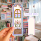 Decorative bookmark with a fairy and butterfly design held by a hand in front of a bookshelf.