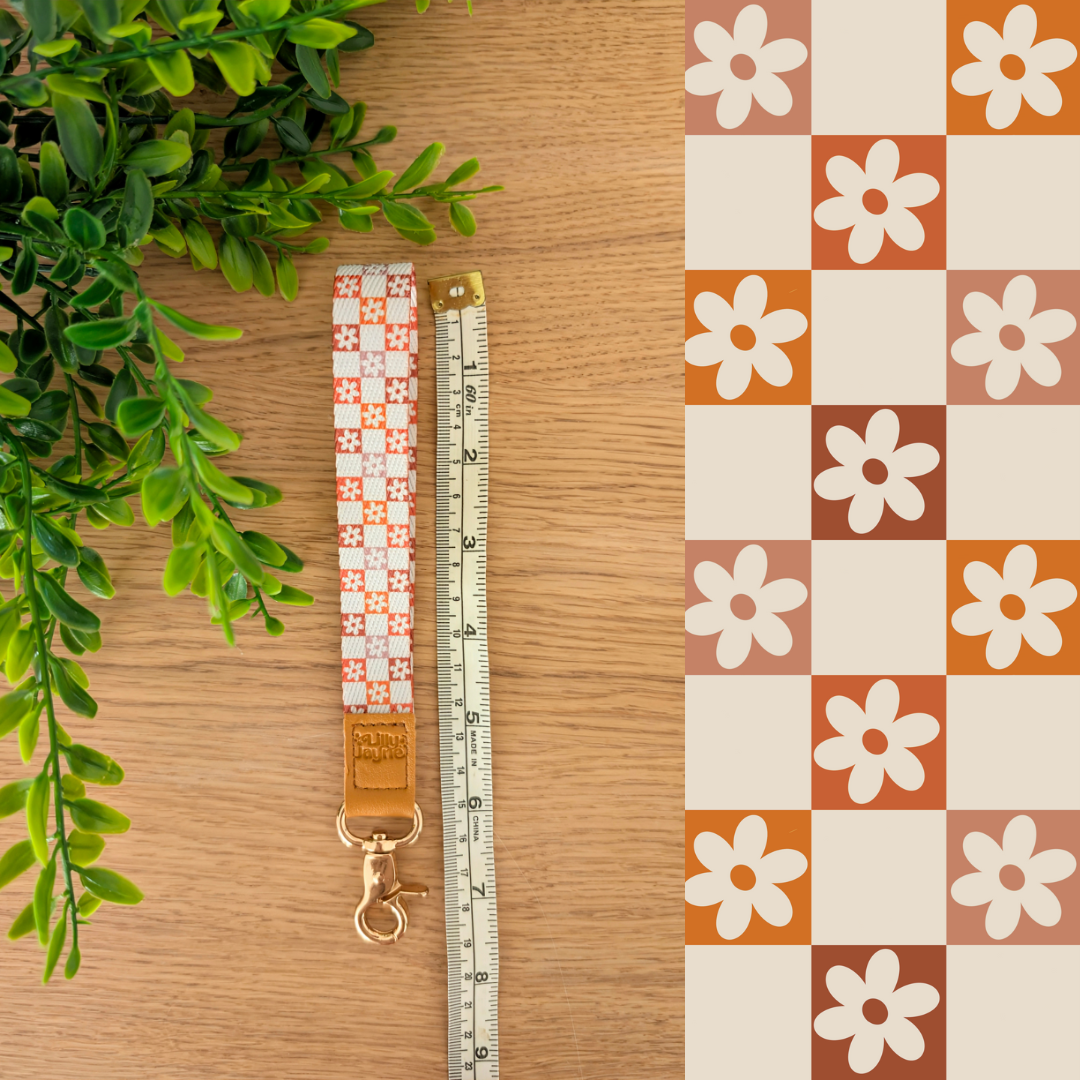 Three colourful fabric wristlet keychains with gold hardware and tan leather ends, featuring pink gingham daisy, blush polka dot stripe, and retro orange checker floral patterns, laid on pastel notebooks with floral accents – Lilly Jayne Collective.