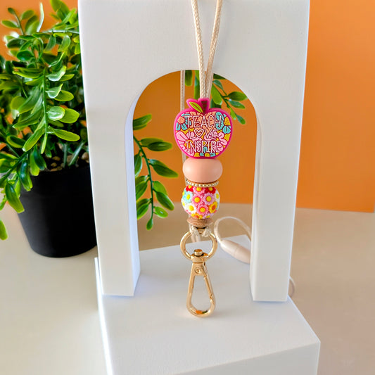 A lanyard featuring a multicolored floral silicone bead design with a gold-colored clasp, displayed against a white stand with a plant in the background.