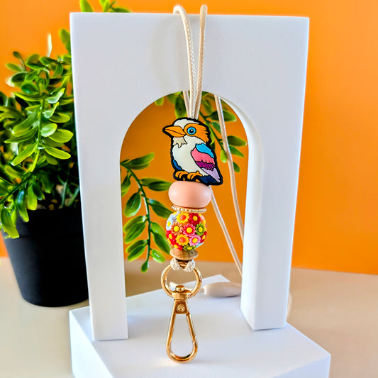 A colorful lanyard featuring a kookaburra design with silicone beads, displayed on a white stand with an orange background and a plant to the side.