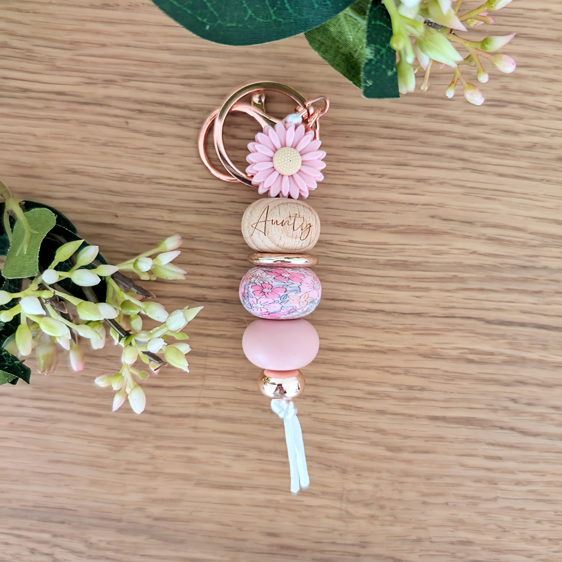 Handmade keyring with pink daisy flower charm, engraved 'Aunty' wooden bead, floral patterned bead, and pastel pink accents on a wooden surface with greenery