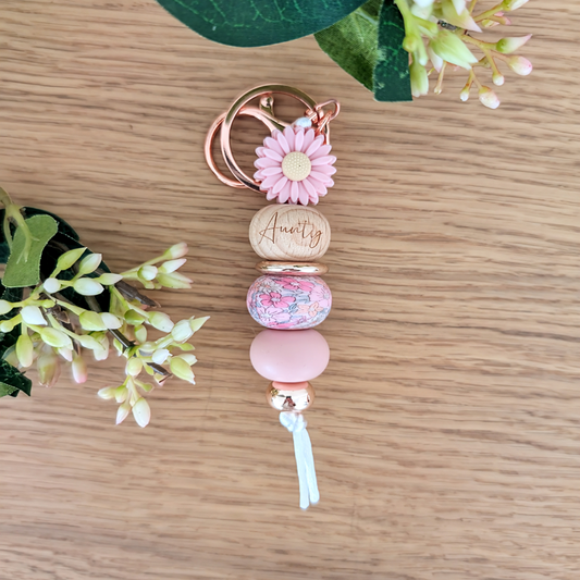 Handmade keyring with pink daisy flower charm, engraved 'Aunty' wooden bead, floral patterned bead, and pastel pink accents on a wooden surface with greenery