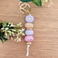 Handmade keyring with purple daisy flower charm, engraved 'Aunty' wooden bead, floral patterned bead, and pastel pink accents on a wooden surface with greenery