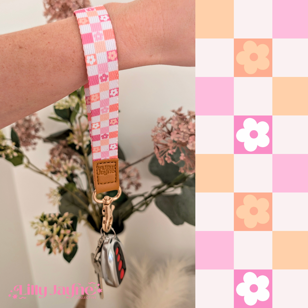 Three colourful fabric wristlet keychains with gold hardware and tan leather ends, featuring pink gingham daisy, blush polka dot stripe, and retro orange checker floral patterns, laid on pastel notebooks with floral accents – Lilly Jayne Collective.