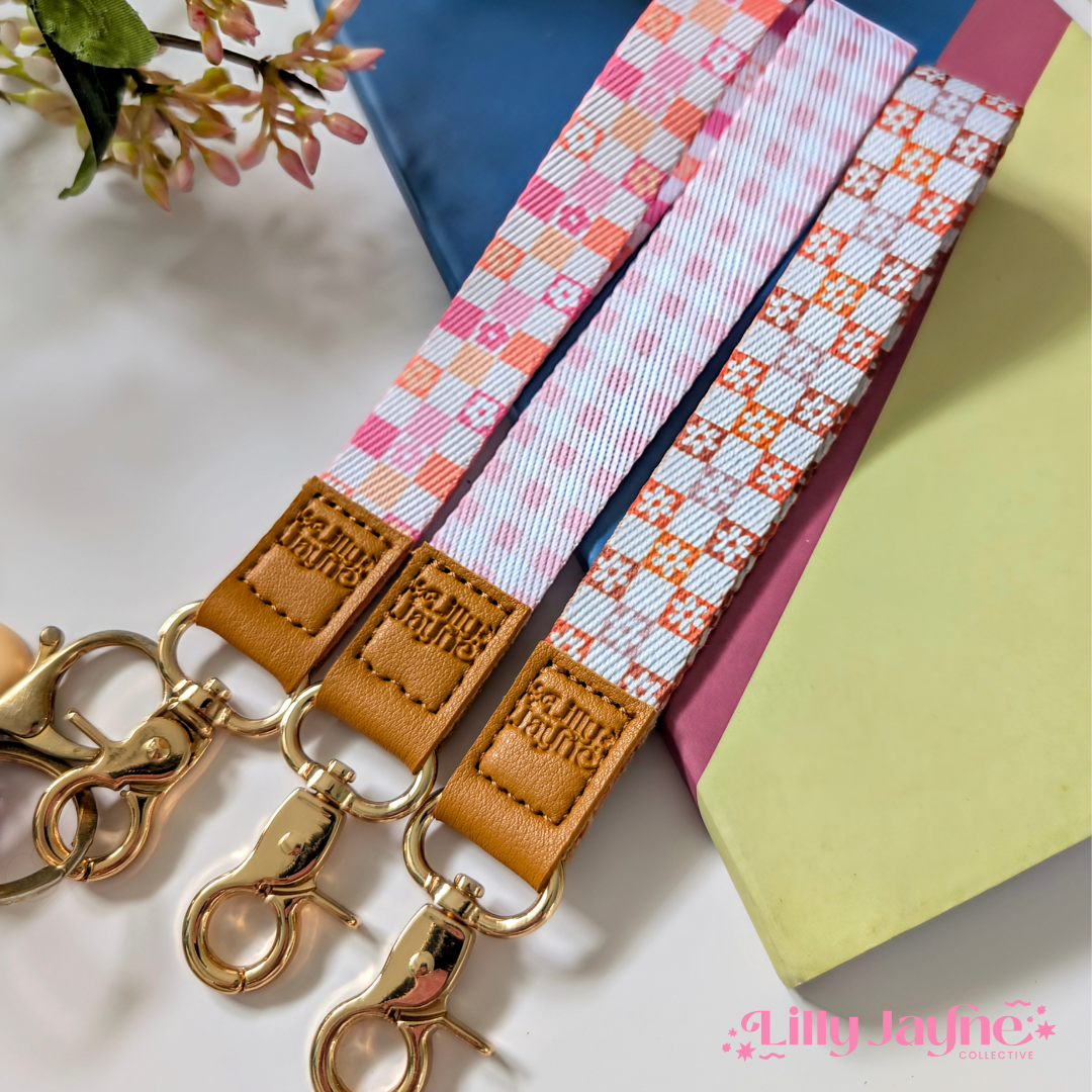 Three colourful fabric wristlet keychains with gold hardware and tan leather ends, featuring pink gingham daisy, blush polka dot stripe, and retro orange checker floral patterns, laid on pastel notebooks with floral accents – Lilly Jayne Collective.