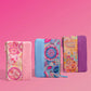 Three colorful zip-around pouches with patterned fabric and solid fabric panels, arranged side by side on a solid pink background.