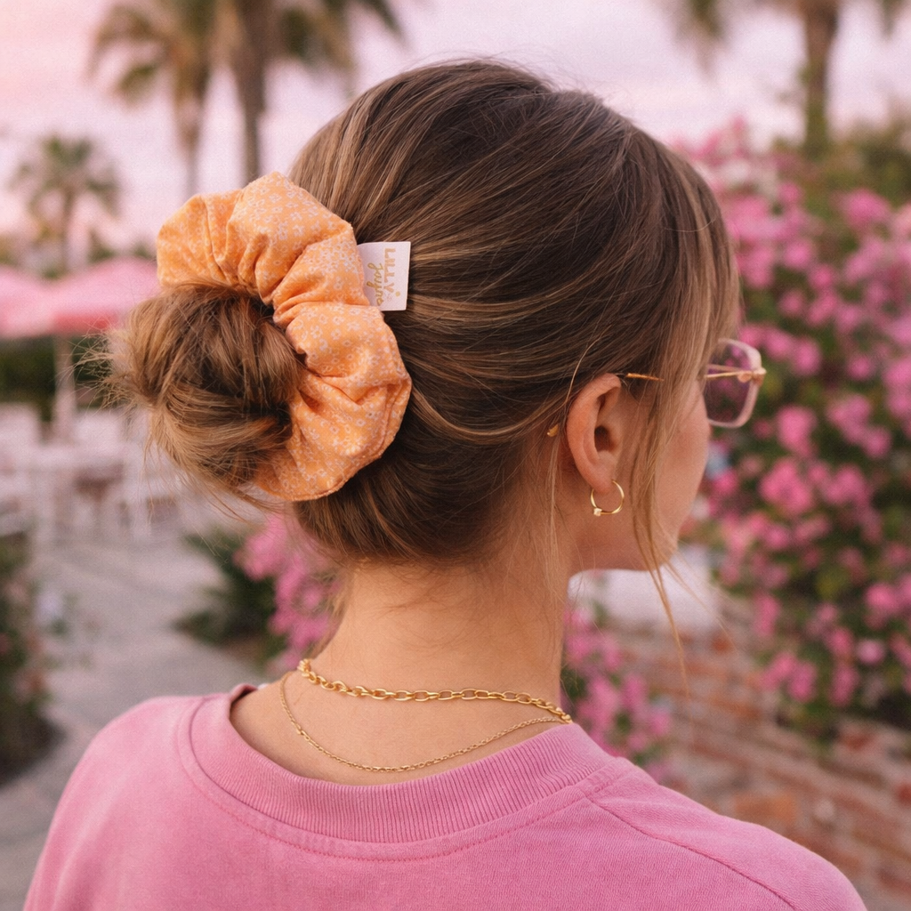 Person with a scrunchie in their hair, wearing a pink shirt and gold jewelry, with a blurred outdoor background.