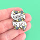 Pin with 'Kindness is Easy' text held between fingers against a green background