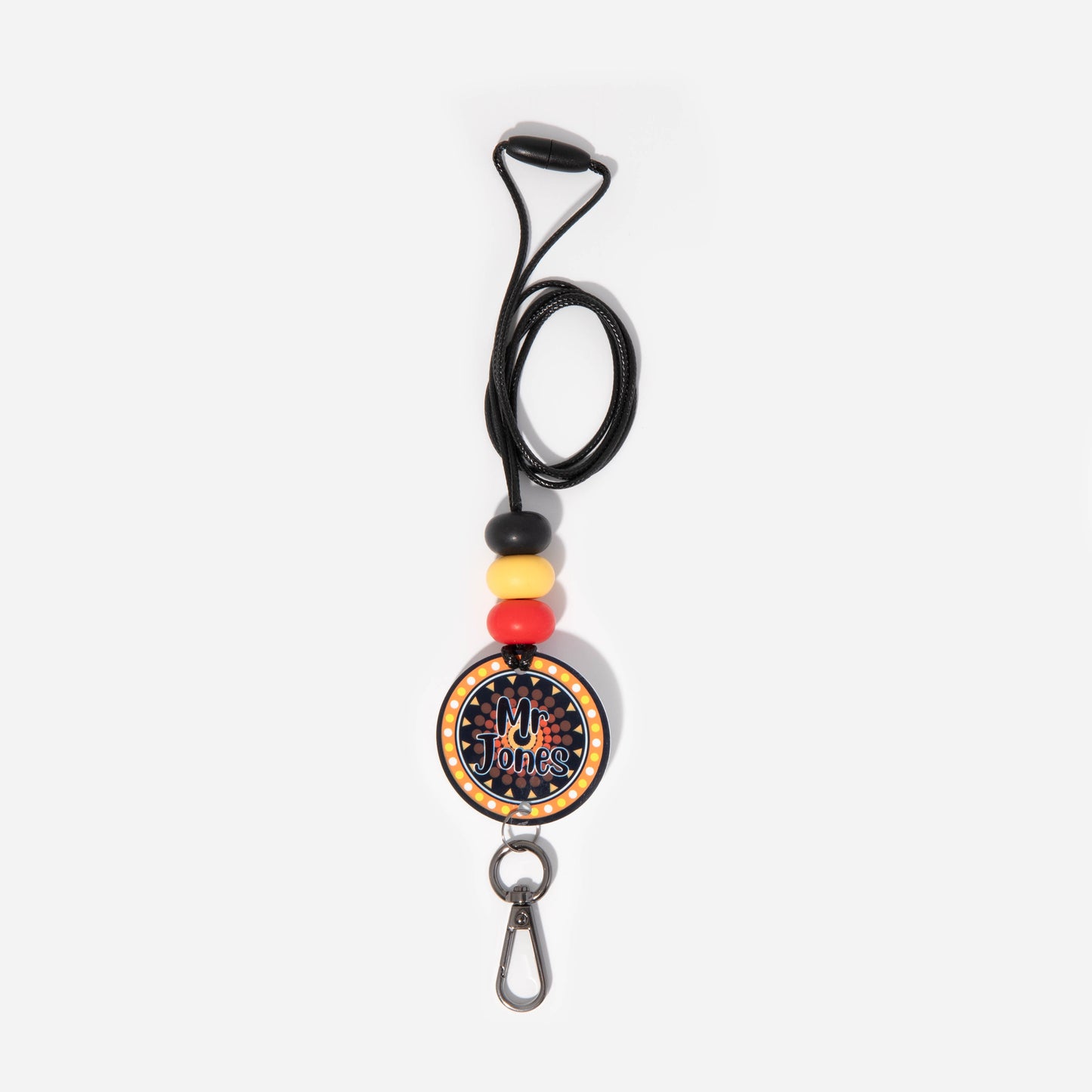 A lanyard featuring a round badge with Aboriginal art patterns, attached to a metal clip and a key ring, with silicone beads in black, red, and yellow colors along the strap.