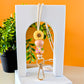 A lanyard with a yellow flower design, featuring a gold clasp and silicone beads, displayed against a white arch with an orange background and a plant to the left.