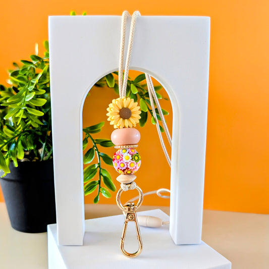 A lanyard with a yellow flower design, featuring a gold clasp and silicone beads, displayed against a white arch with an orange background and a plant to the left.