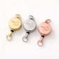 Three retractable keychains in gold, silver, and rose gold on a white background.