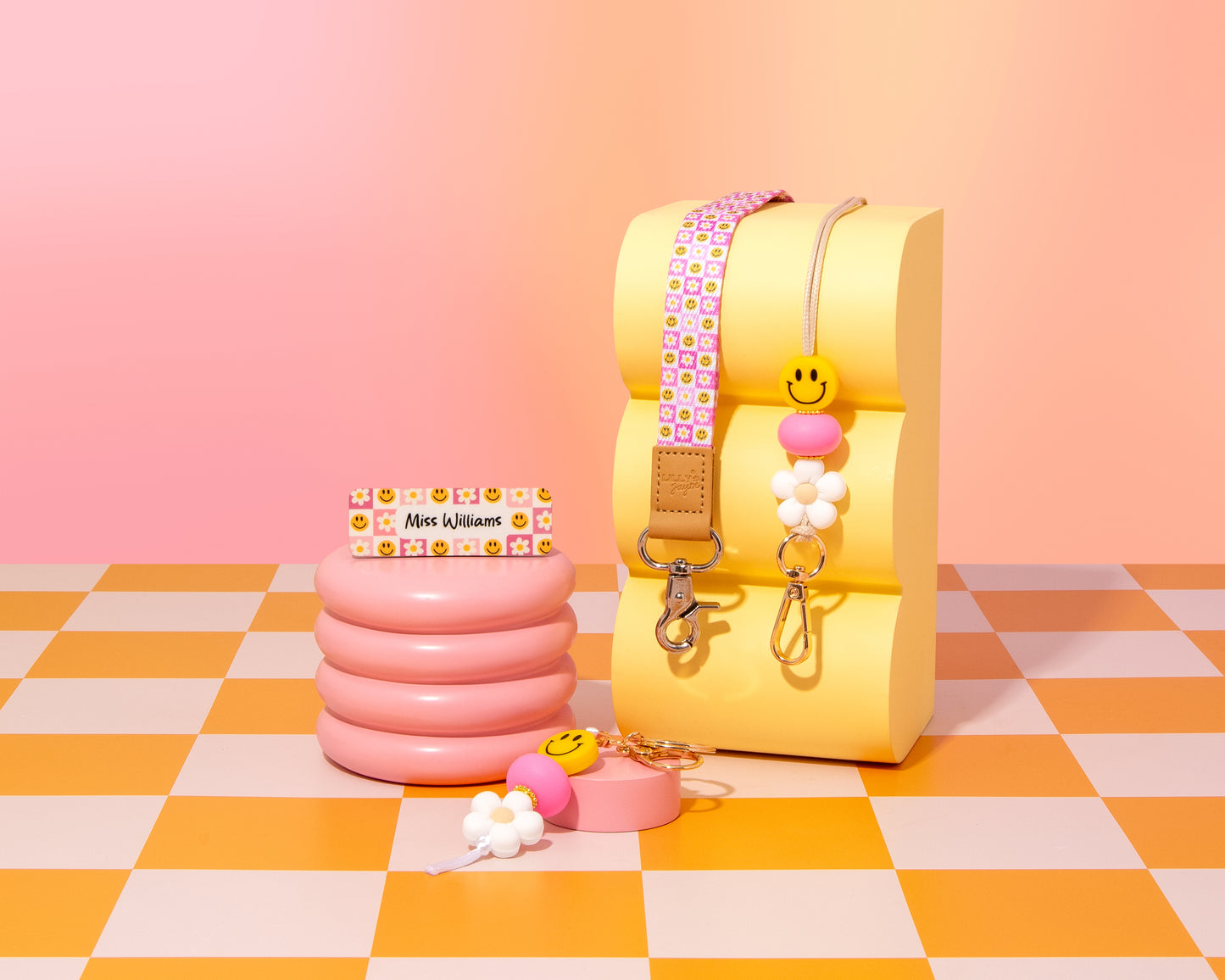 Colorful children's products including a yellow seat, pink inflatable cup holder, and keychains on a checkered floor with pink background.