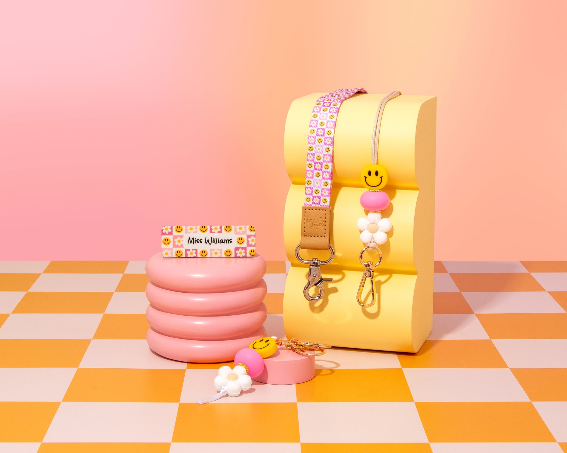 Colorful children's products including a yellow seat, pink inflatable cup holder, and keychains on a checkered floor with pink background.