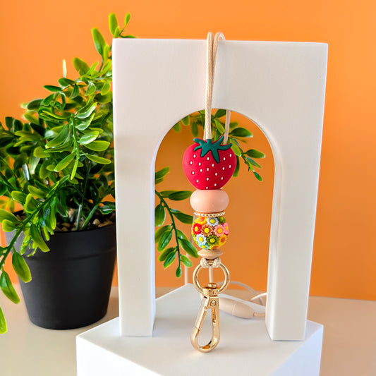 A lanyard with a pink flower design, featuring a silicone bead, attached to a safety clasp. Displayed on a white stand with a plant in the background.
