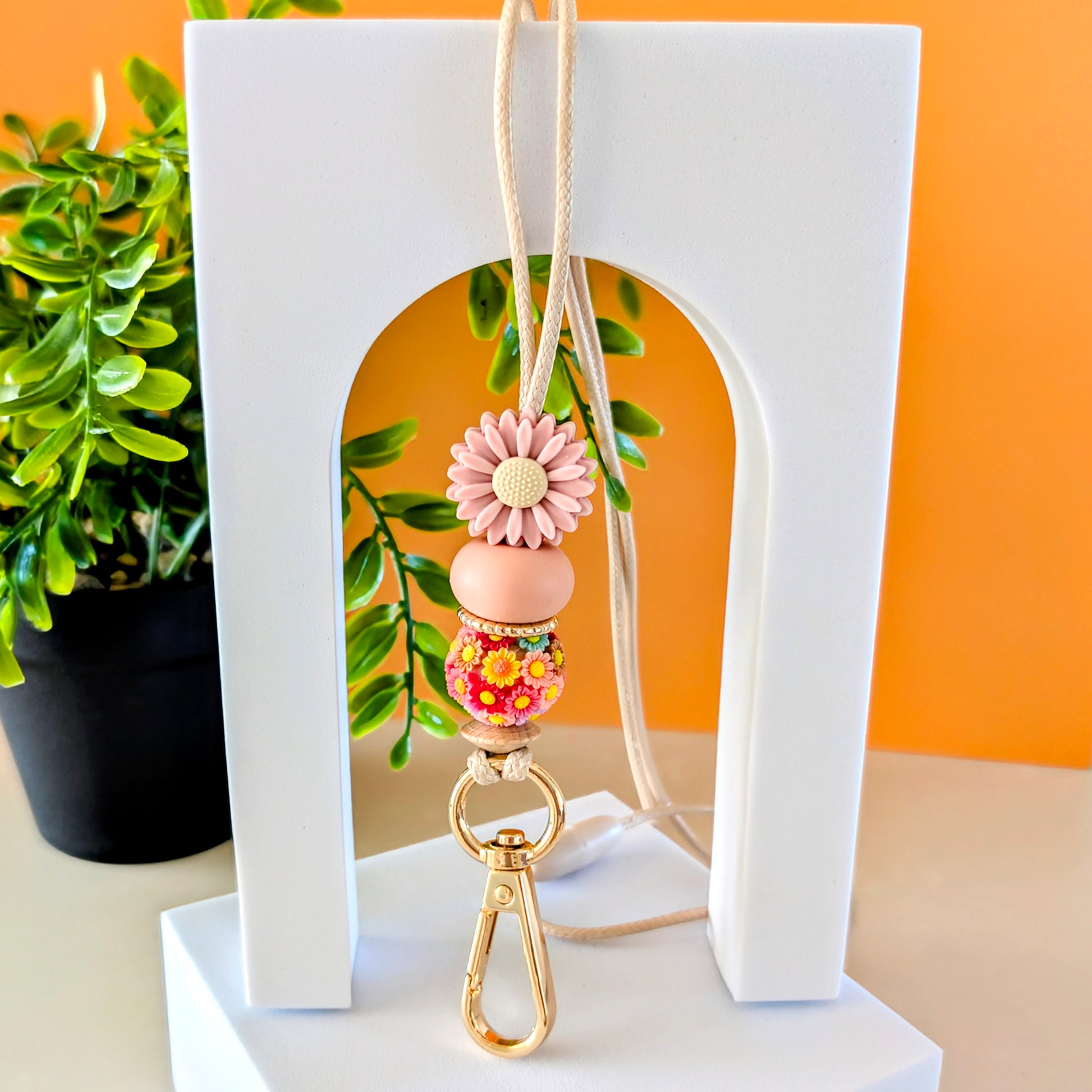 A pink flower lanyard with silicone beads, designed to hold keys or ID cards, displayed on a white stand with a plant in the background.